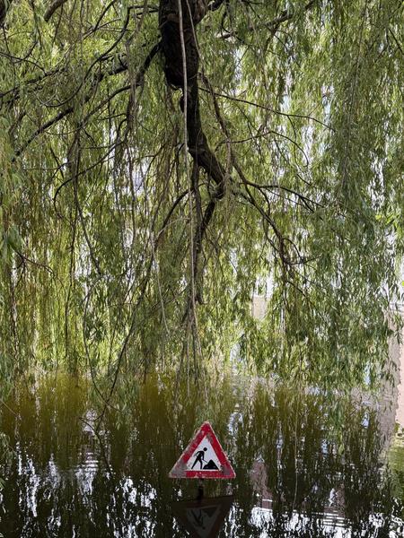 A striking image captured at Kattenburgerkade, Kattenburg, Amsterdam, shows a 'men at work' traffic sign partially submerged in dark water, reflecting the lush green canopy of a weeping willow tree overhead